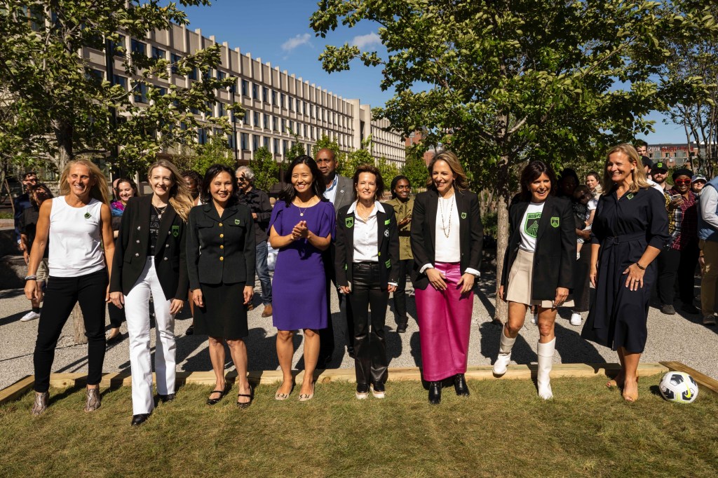 Eight women stand on grass with a soccer ball on the right hand side of the grass. Many of the women wear matching black jackets with the Boston Unity Soccer Club green logo
